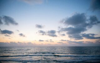 A picture from the shore of the beach as the sun it setting over the ocean waters. There are speckled clouds in the sky.