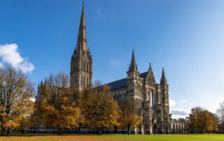 A picture of a towering cathedral with tall, triangular spires reaching to the bright blue sky above. There is a field of grass and autumn trees surrounding it.