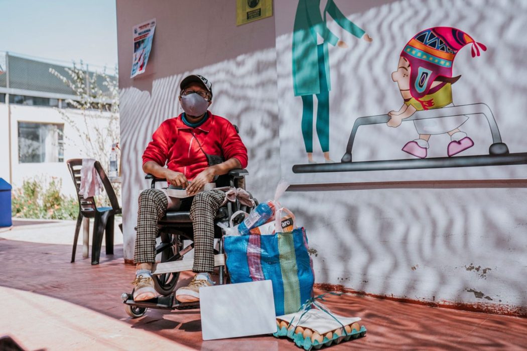 A woman using a wheelchair with a COVID mask on, bags full of groceries, an egg carton, and a colorful mural behind her.
