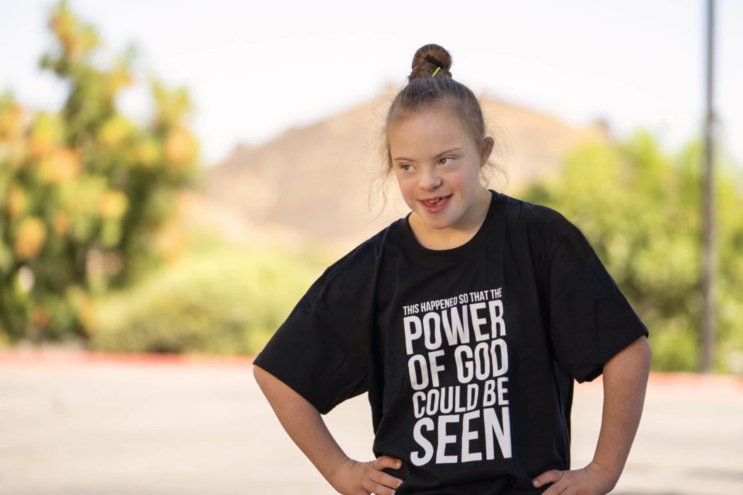 A young girl who appears to have down syndrome with a shirt on that says "This happened so that the power of God could be seen."