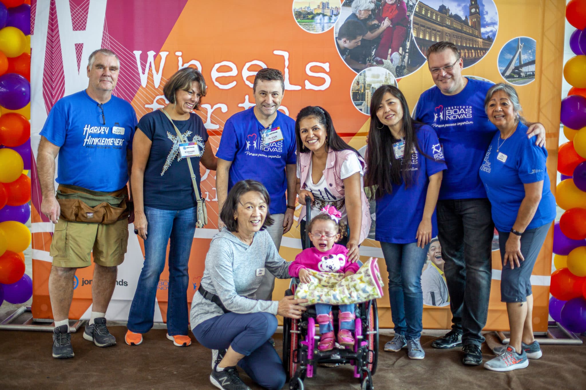 A photo of a family of people surrounding a young girl using a small wheelchair with a bow in her hair and glasses on.