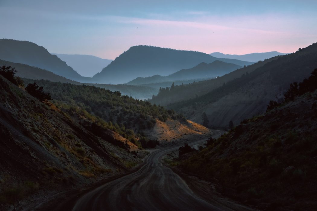 A winding dirt road running through a forested mountain range with the sun setting in the background.