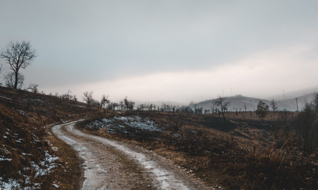 A scorched landscape with a small road running through it and thick clouds above.