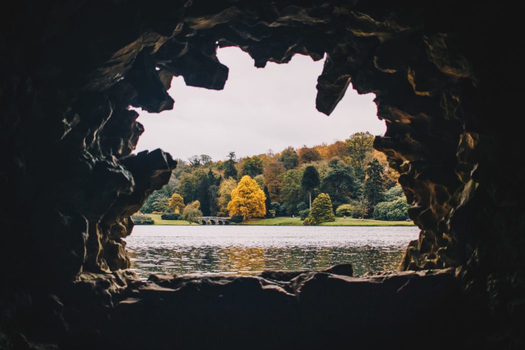 The view through the tunnel of a cave looking out at a lake with a bridge and a thick forest on the other side.