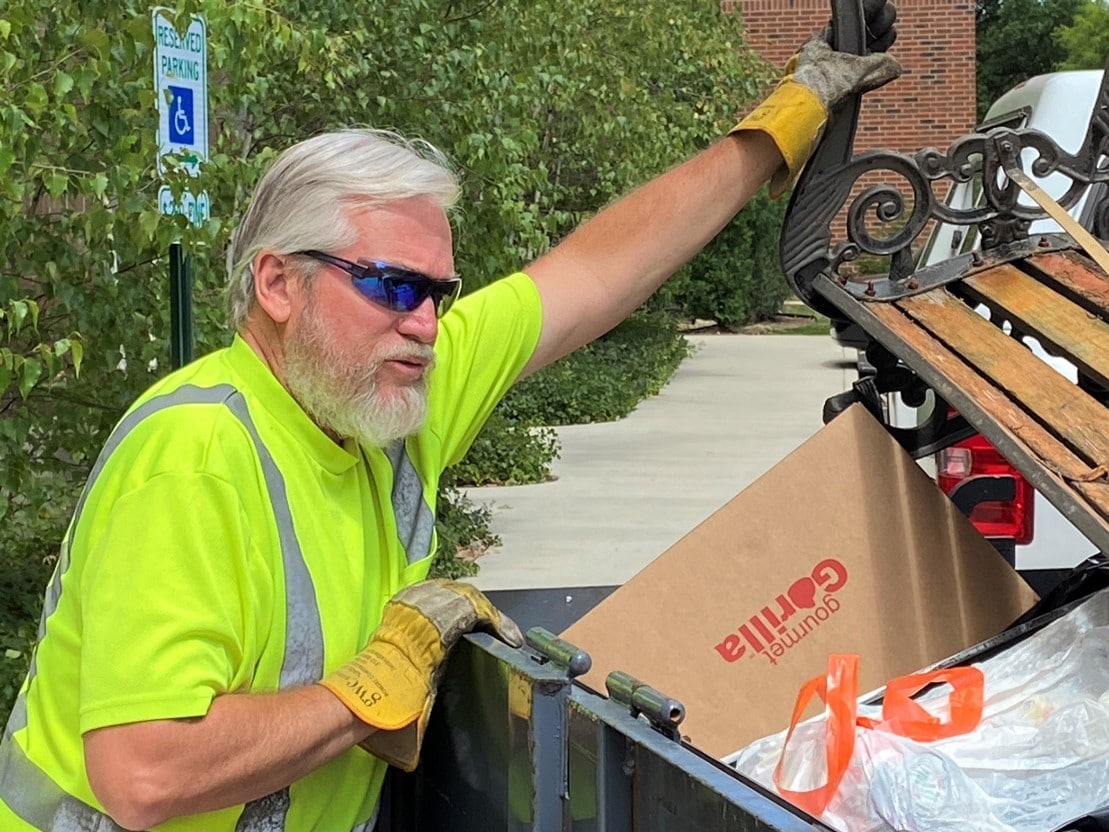 Close up of a man in a construction outfit with sunglasses on as he puts a broken bench in a large trash bin.