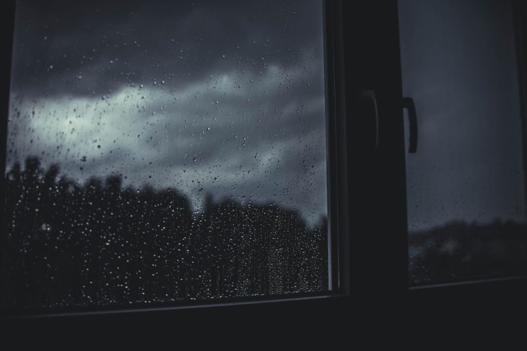 A window with raindrops streaking down with a view of dark storm clouds and a line of trees outside.