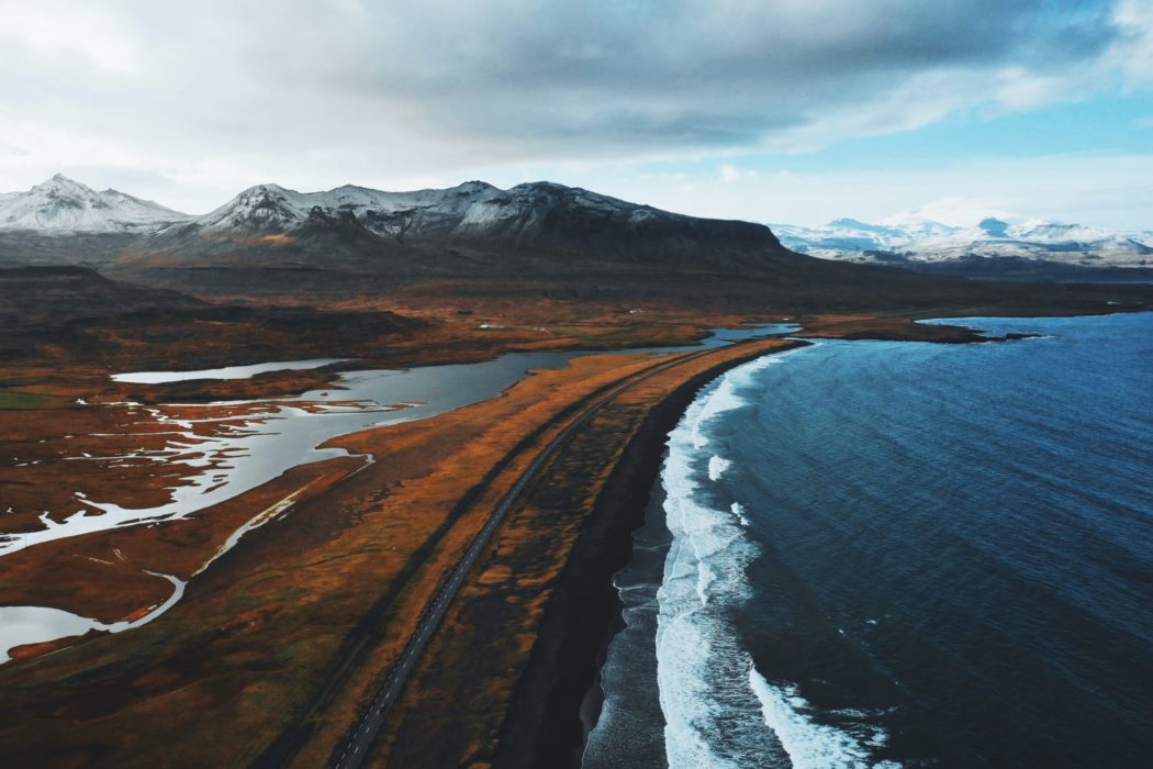 Snowy mountains overlooking the ocean with a two lane road separating the water from the shore.
