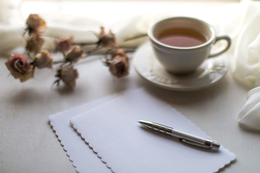 A paper on a table with a pen on it, a tea cup and dried roses next to it.