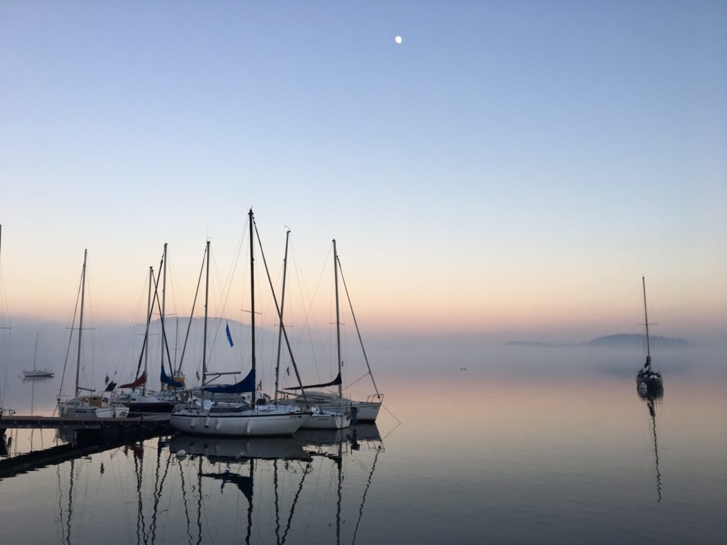 A group of six sail boats docked at a harbor as the sunset reflects off the water.