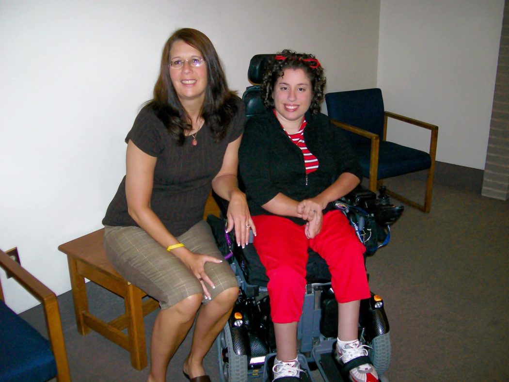 A mom sitting next to her daughter who is using a wheelchair as they both smile at the camera.