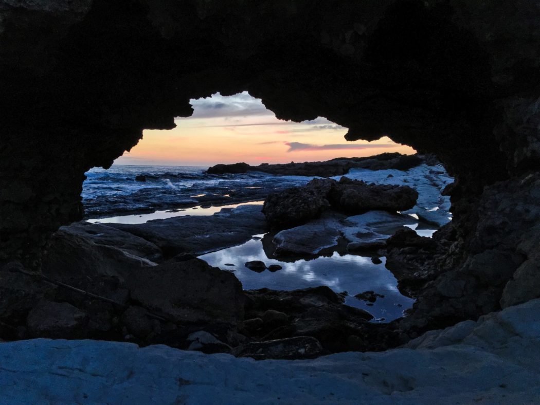 The view looking through a rocky tunnel at the beach where there is rocks all along the beach with the waves crashing on the shore.