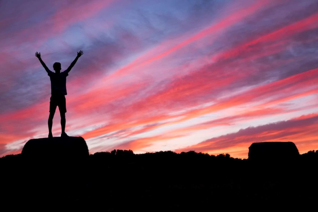 A man standing on a rock with his arms raised in the air looking at a sunset sky.