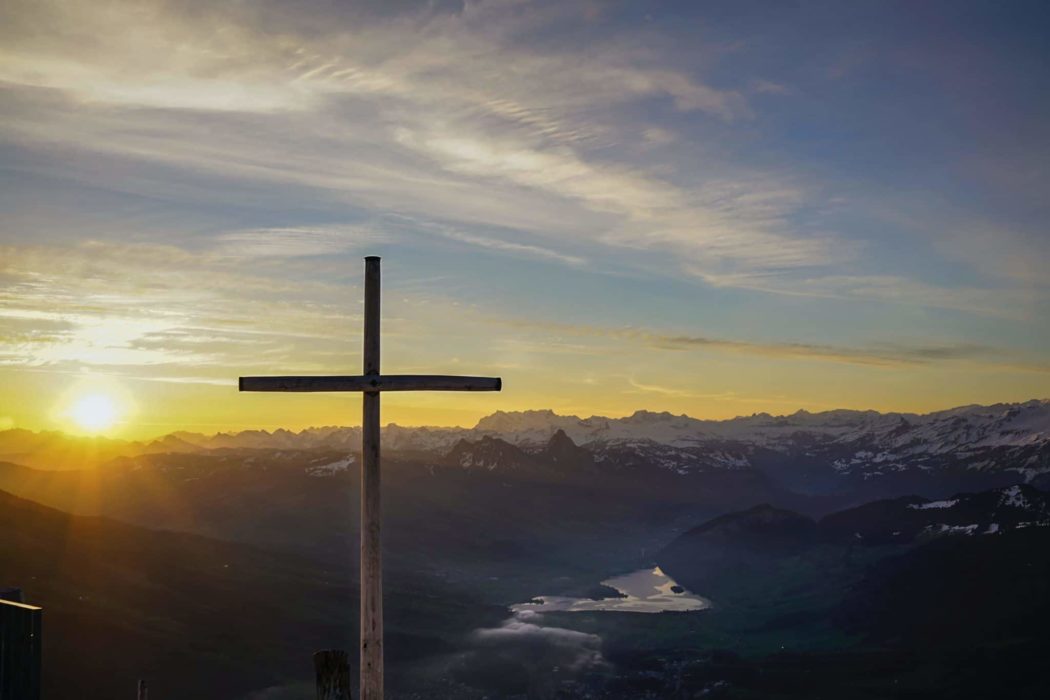 A cross with a sunset sky and a mountain range behind it.