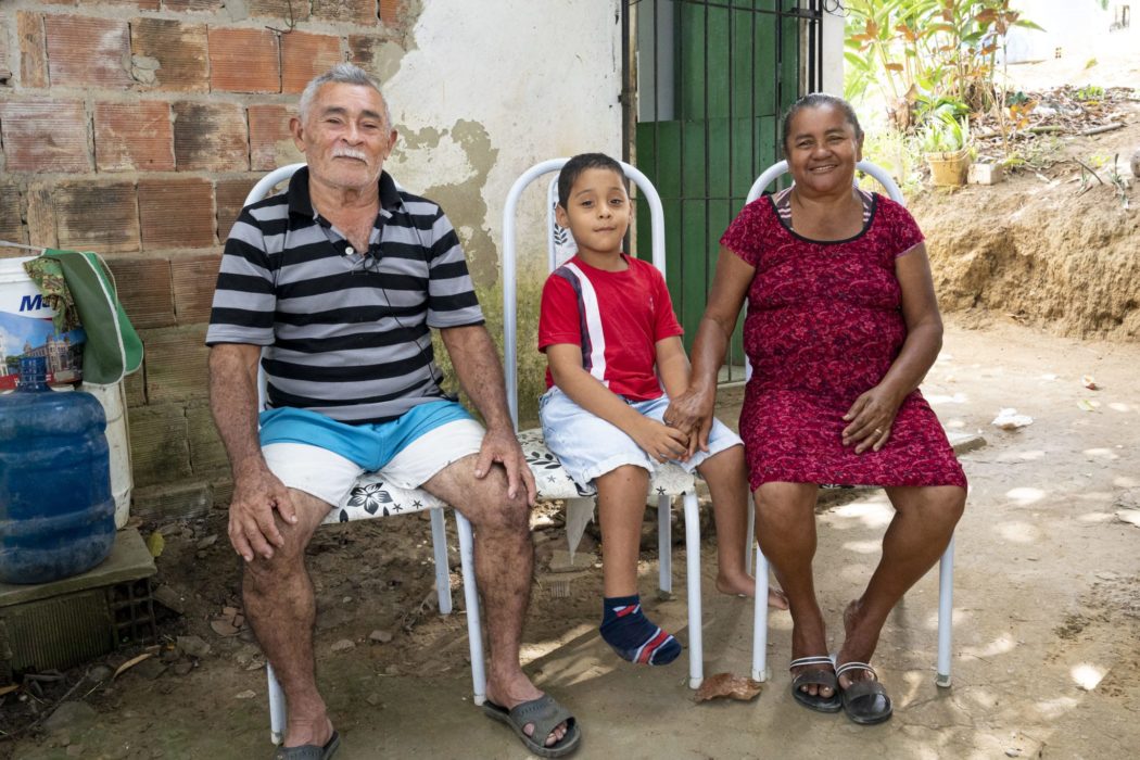 A family including a man, a woman and a young boy, sitting outside of a brick building in upright chairs smiling at the camera.
