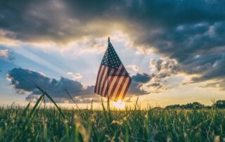 A small American flag standing in the middle of a field of grass as the sun is setting behind it. Grey, scattered clouds are in the sky above.