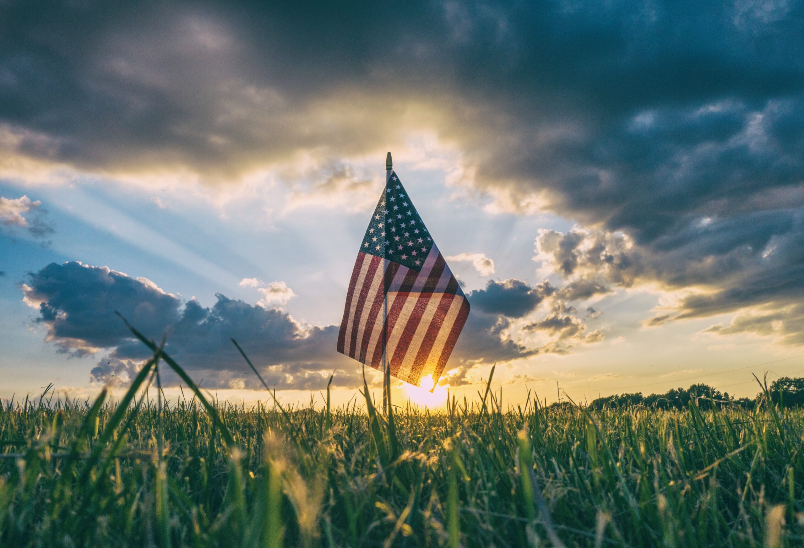 A small American flag standing in the middle of a field of grass as the sun is setting behind it. Grey, scattered clouds are in the sky above.