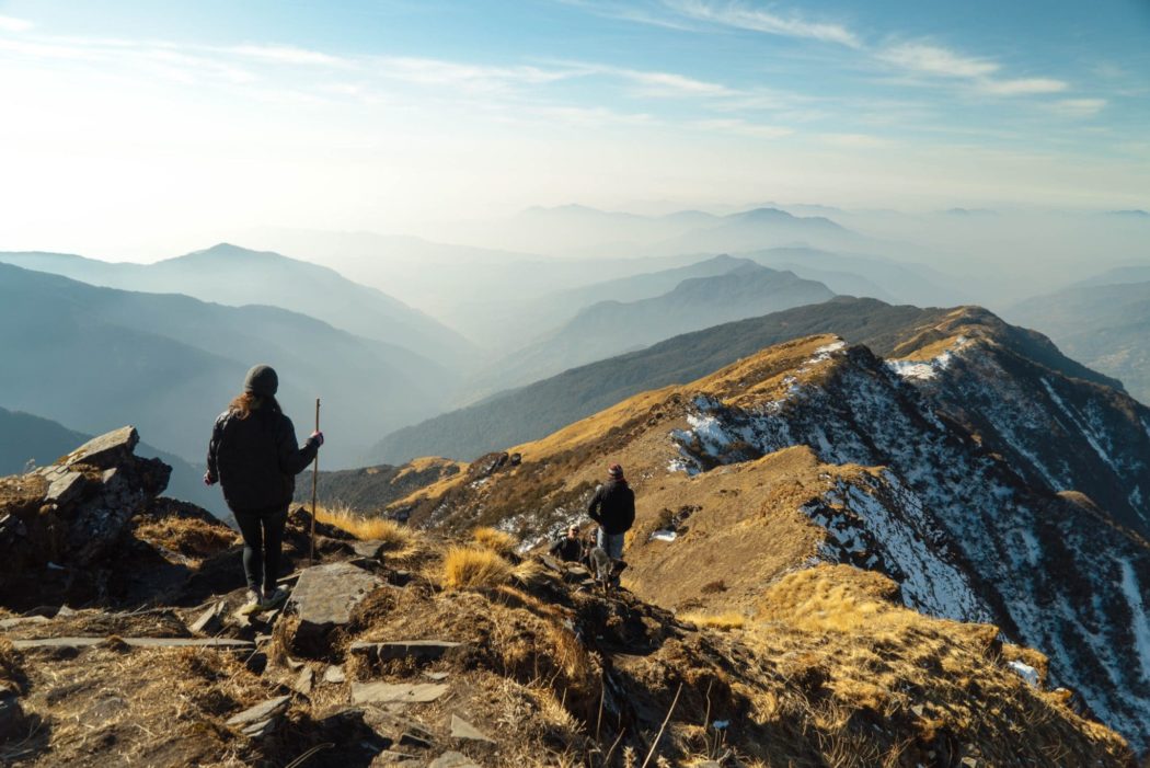 A man and a woman hiking with their dog on the ridge of a mountain overlooking a mountain range that spans for miles.
