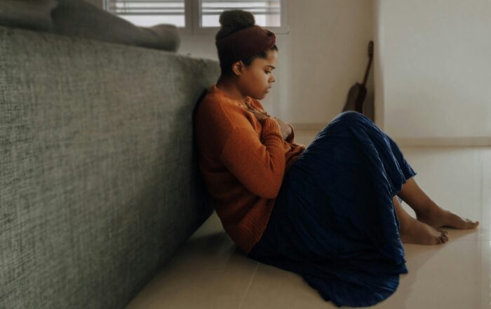 A woman sitting on the floor holding her chest as she looks at the ground with a look of fear on her face.