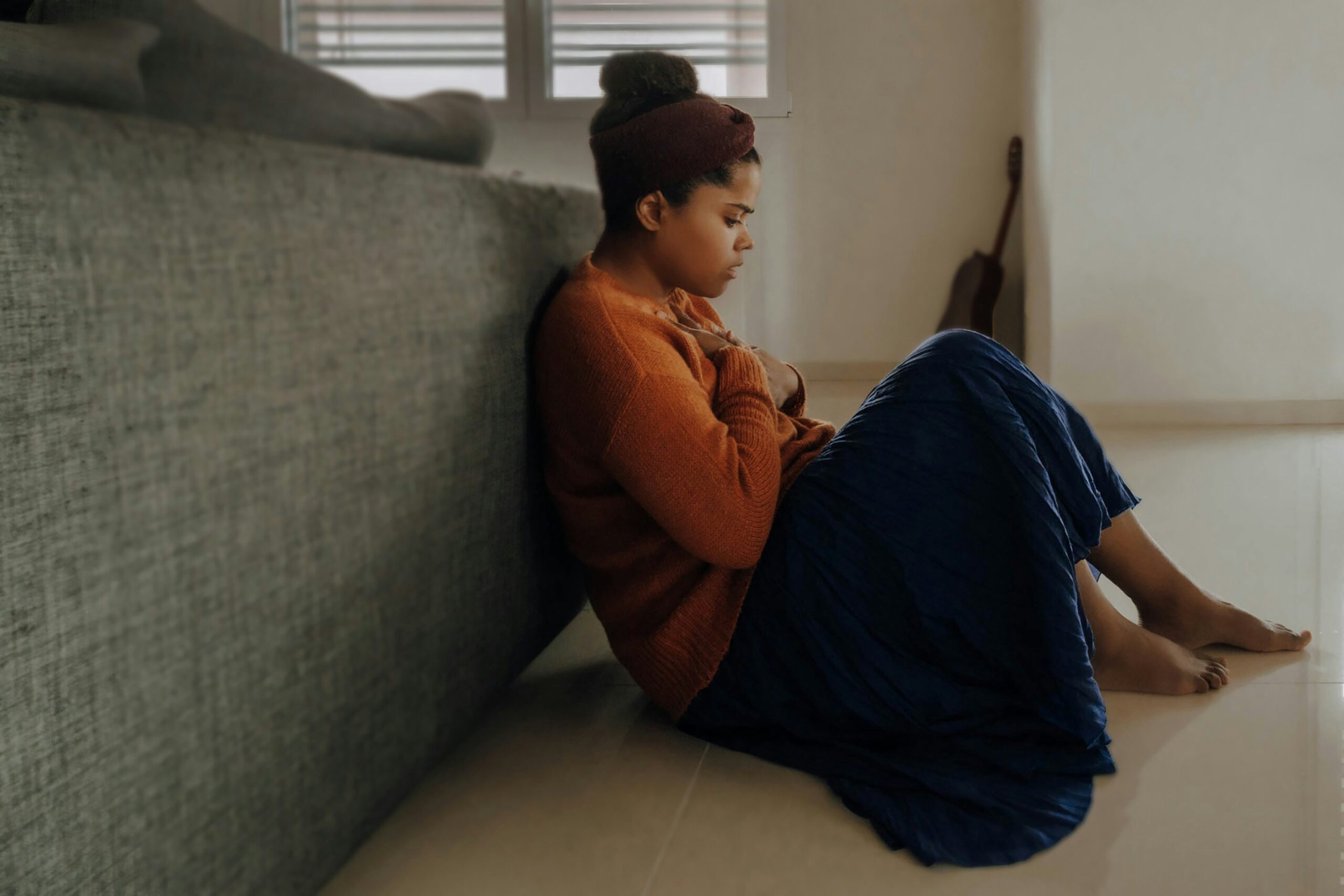 A woman sitting on the floor holding her chest as she looks at the ground with a look of fear on her face.