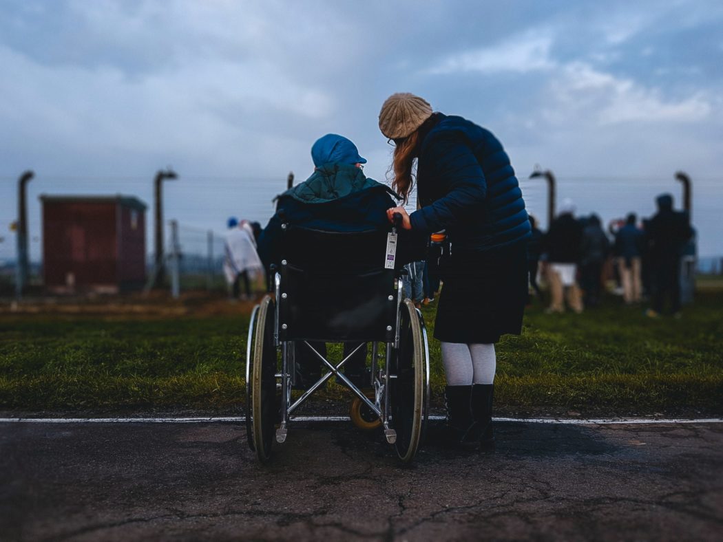 A women leaning close to a man in a wheelchair looking at a barbwire fence.