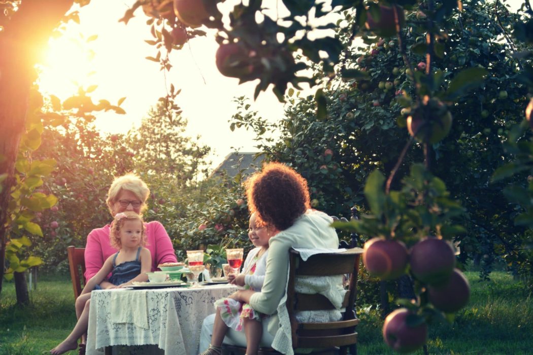 Two older women with young girls sitting at a table laughing in the middle of an apple orchard.