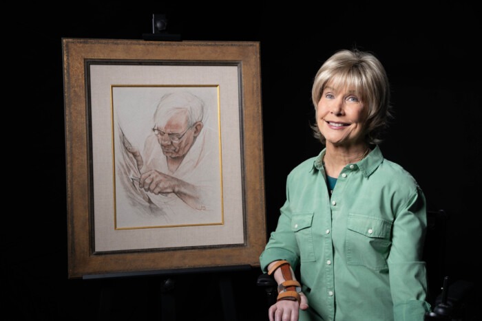 A picture of Joni smiling and sitting next to her drawing of her Father, titled, "My Father's Creation".