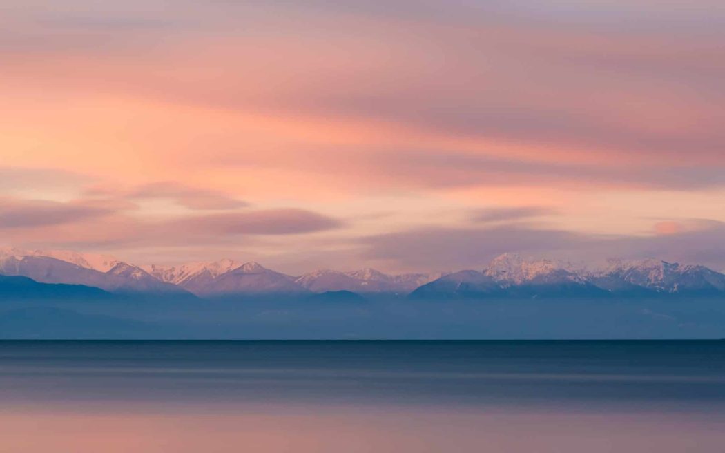 The view from across a body of water with mountains in the background, snow on the peaks, a beautiful sunset overhead.