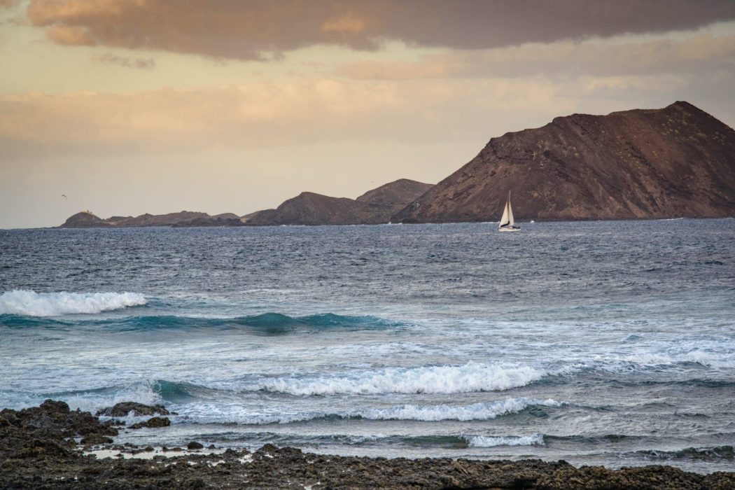 The view from the shore of a sailboat in the distance with an large mountain above it in the background, dark clouds above.