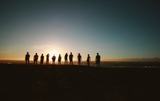 Silhouette of a group of friends standing together on a beach at sunset, with the sun low on the horizon in front of them.
