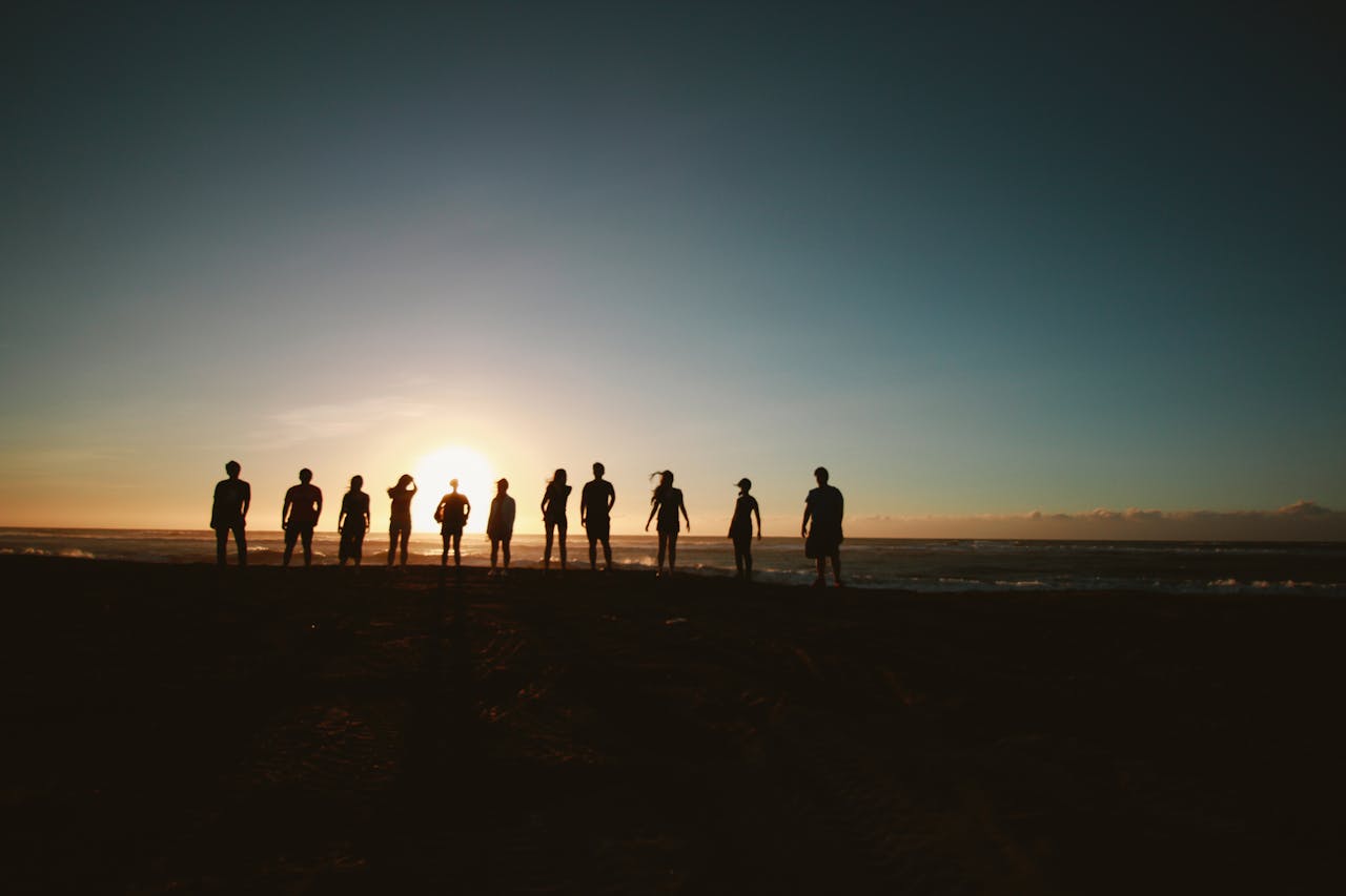 Silhouette of a group of friends standing together on a beach at sunset, with the sun low on the horizon in front of them.