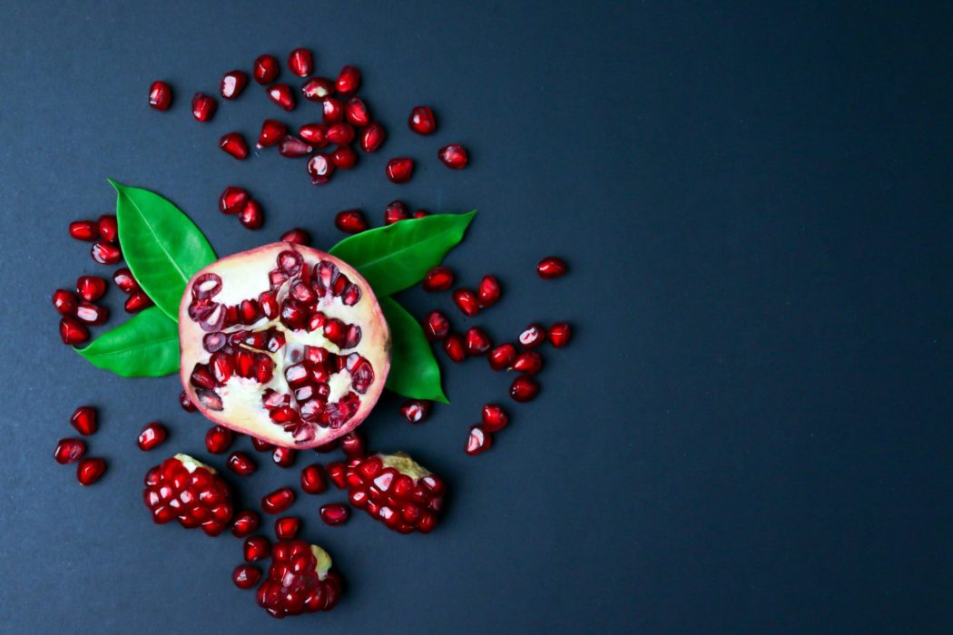  A halved pomegranate with scattered seeds and green leaves arranged on a dark blue background, creating a vibrant contrast between the rich red fruit, bright green leaves, and dark surface.