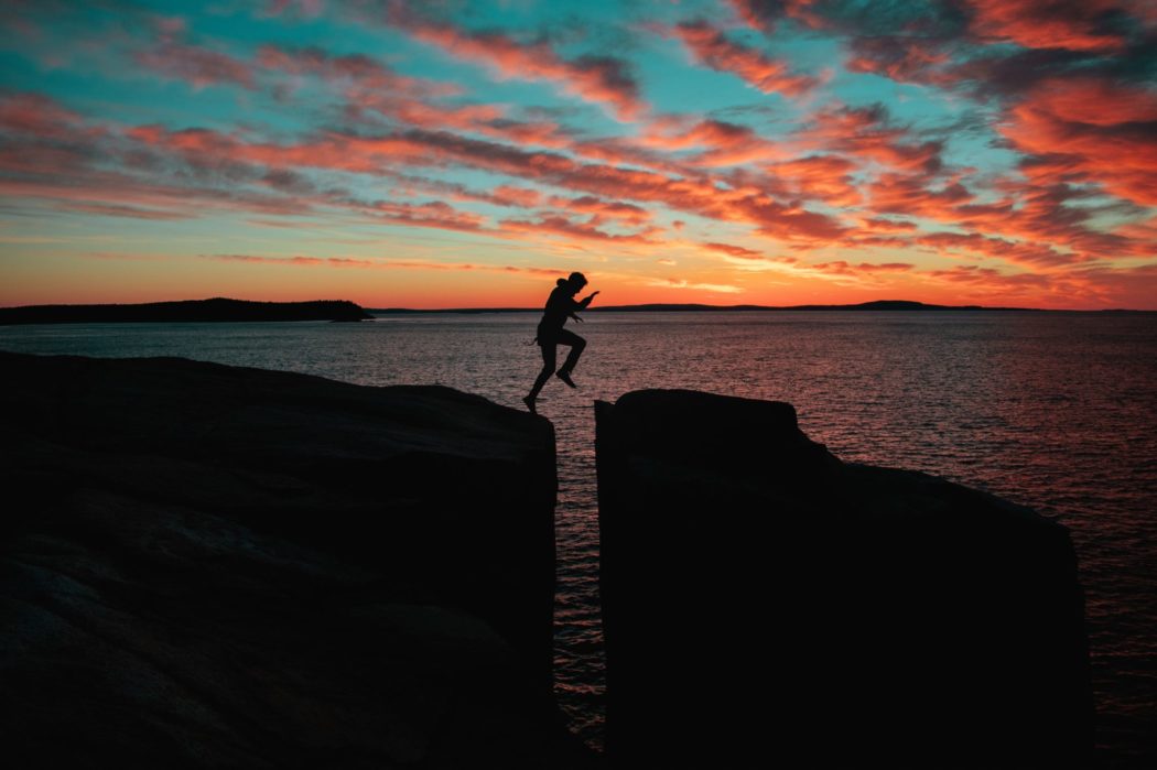 A person in silhouette mid-air, jumping across a gap between two rock formations against a dramatic sunset or sunrise sky. 