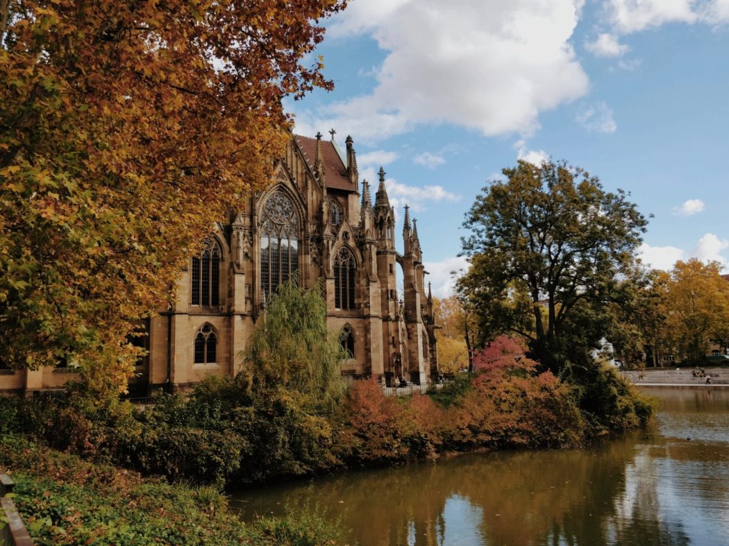 An old church building with a river in the foreground and trees surrounding it.