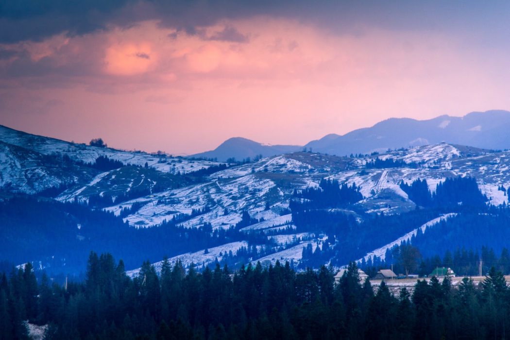 A mountain range with snow-covered peaks and evergreen trees. The sky has a pink and purple hue, suggesting either sunrise or sunset.