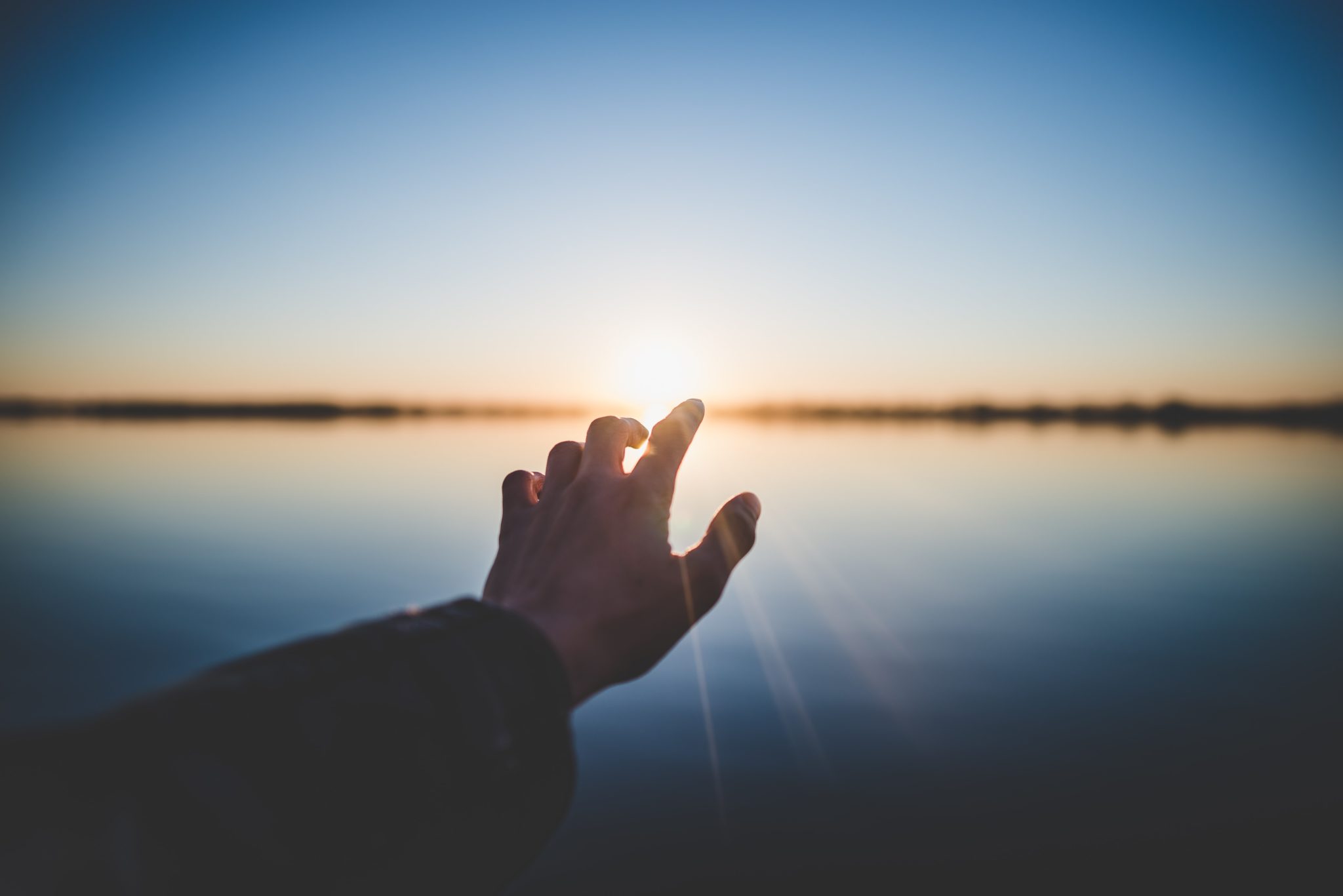 Close up of a man's hand reaching out for the sunrise on the horizon.