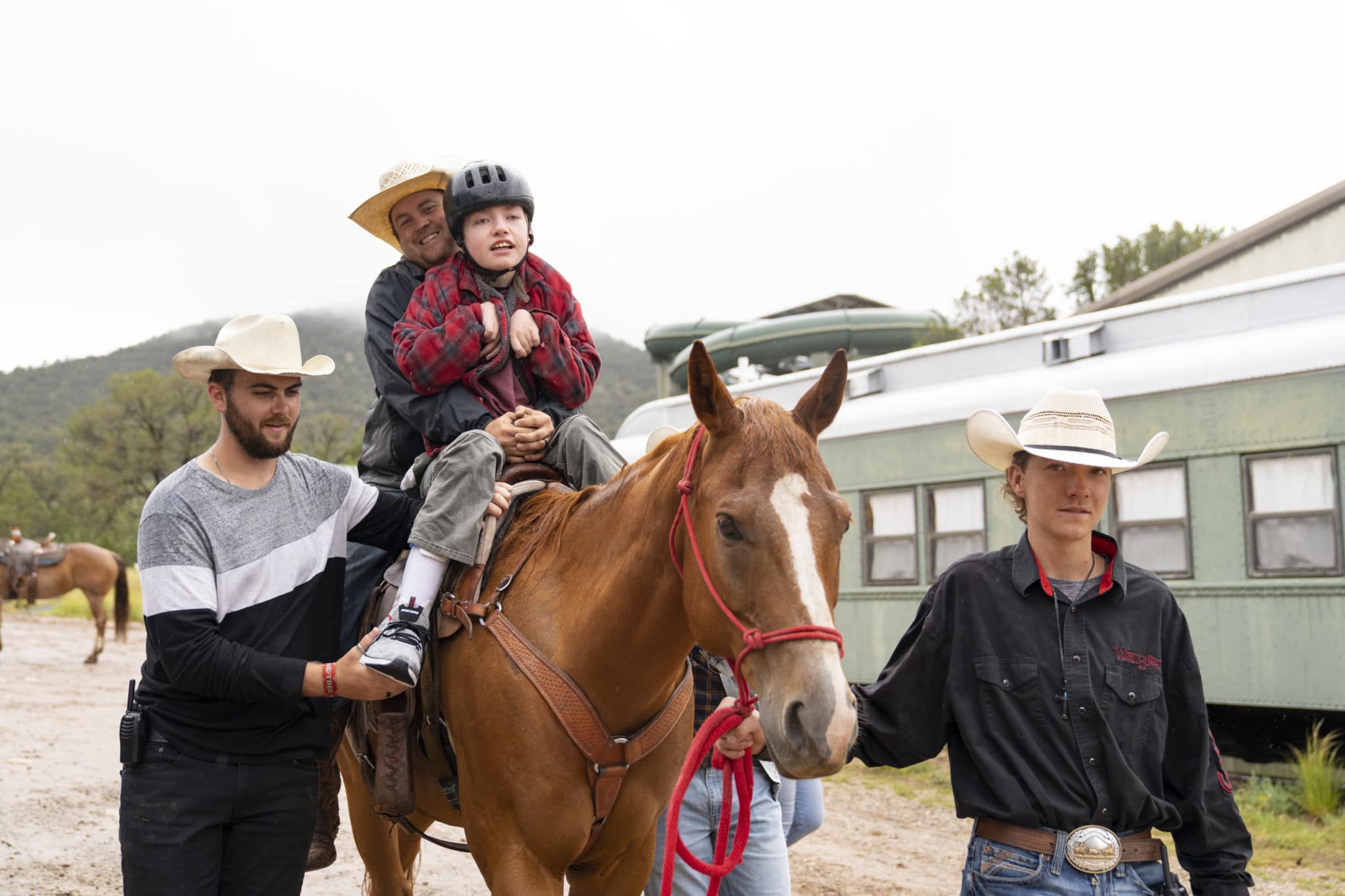Tyler is out of his wheelchair and riding on the back of a horse with a Family Retreat volunteer. Two other volunteers stand on either side of the horse.