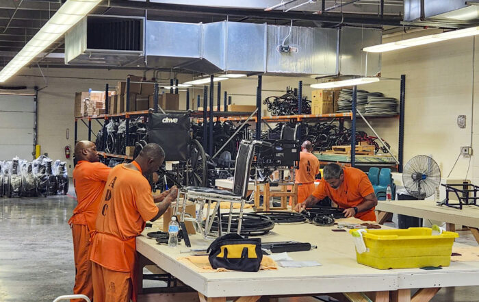 Inmates wearing bright orange uniforms concentrate on repairing wheelchairs in a workshop setting.