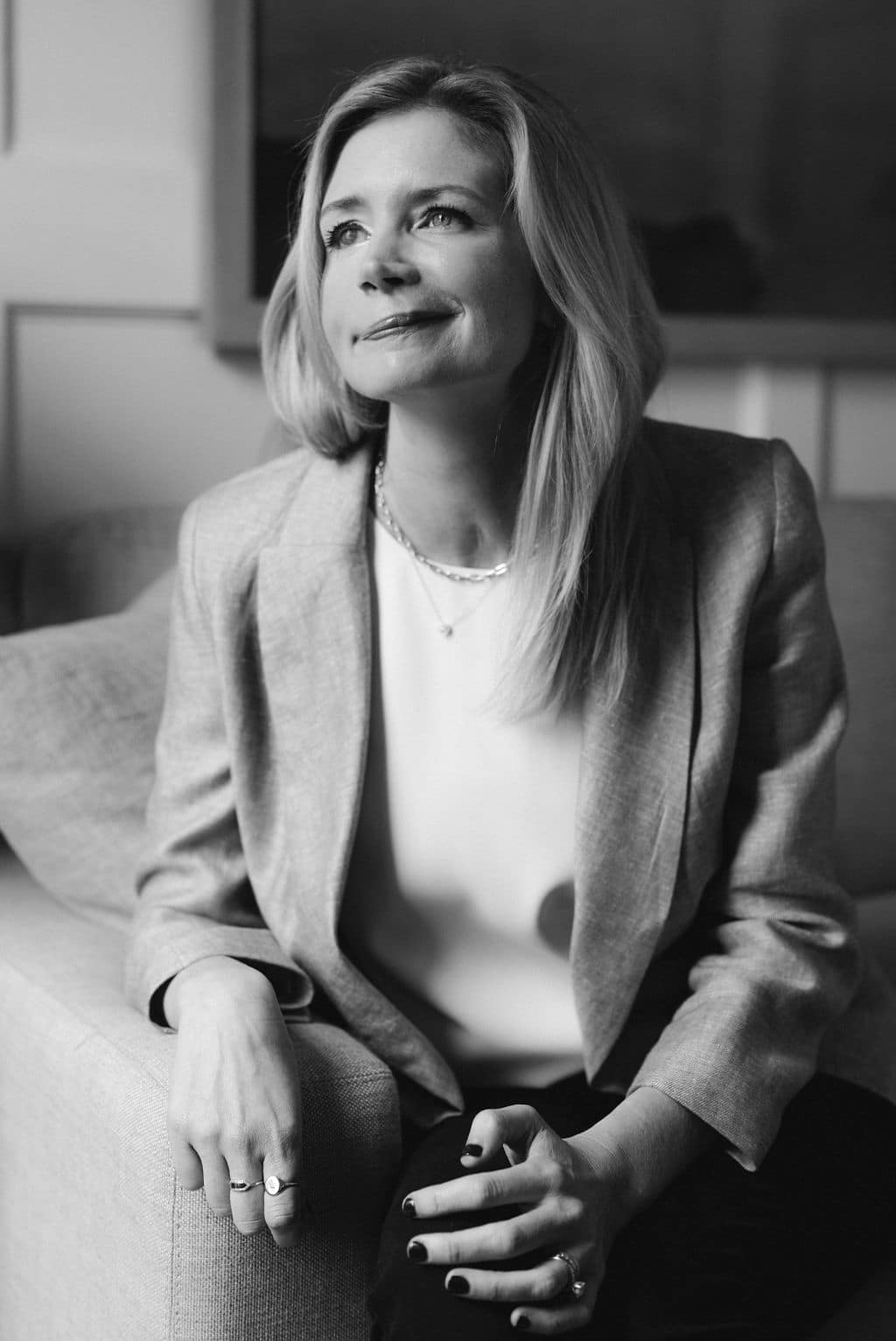 Katherine Wolf sitting on the end of a couch, her left hand over the arm of the couch, her other hand resting on her left knee. She smiles as she looks away from the camera into the distance.