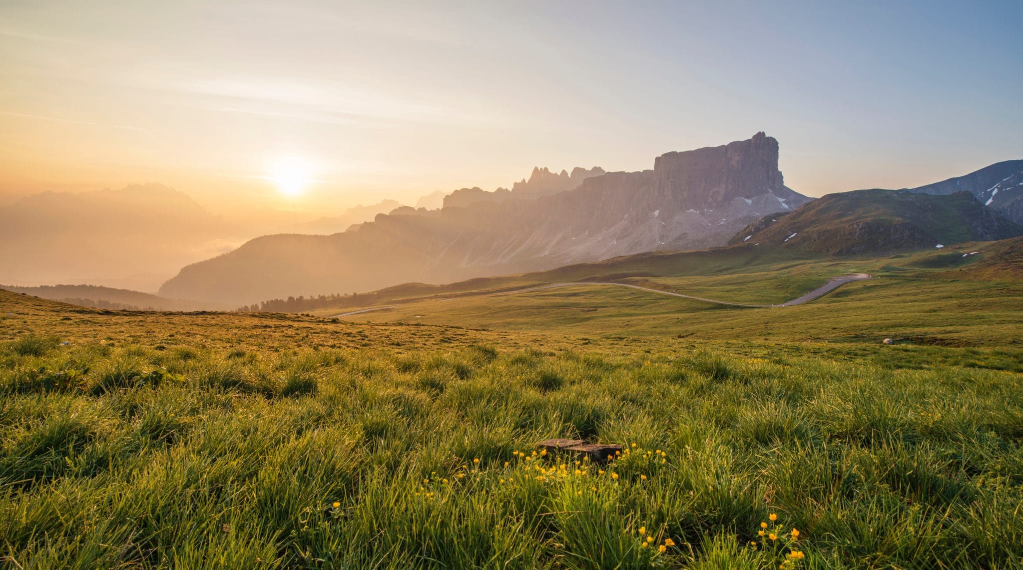 The setting sun beams through clouds across a meadow landscape, cascading over a mountain range in the background.