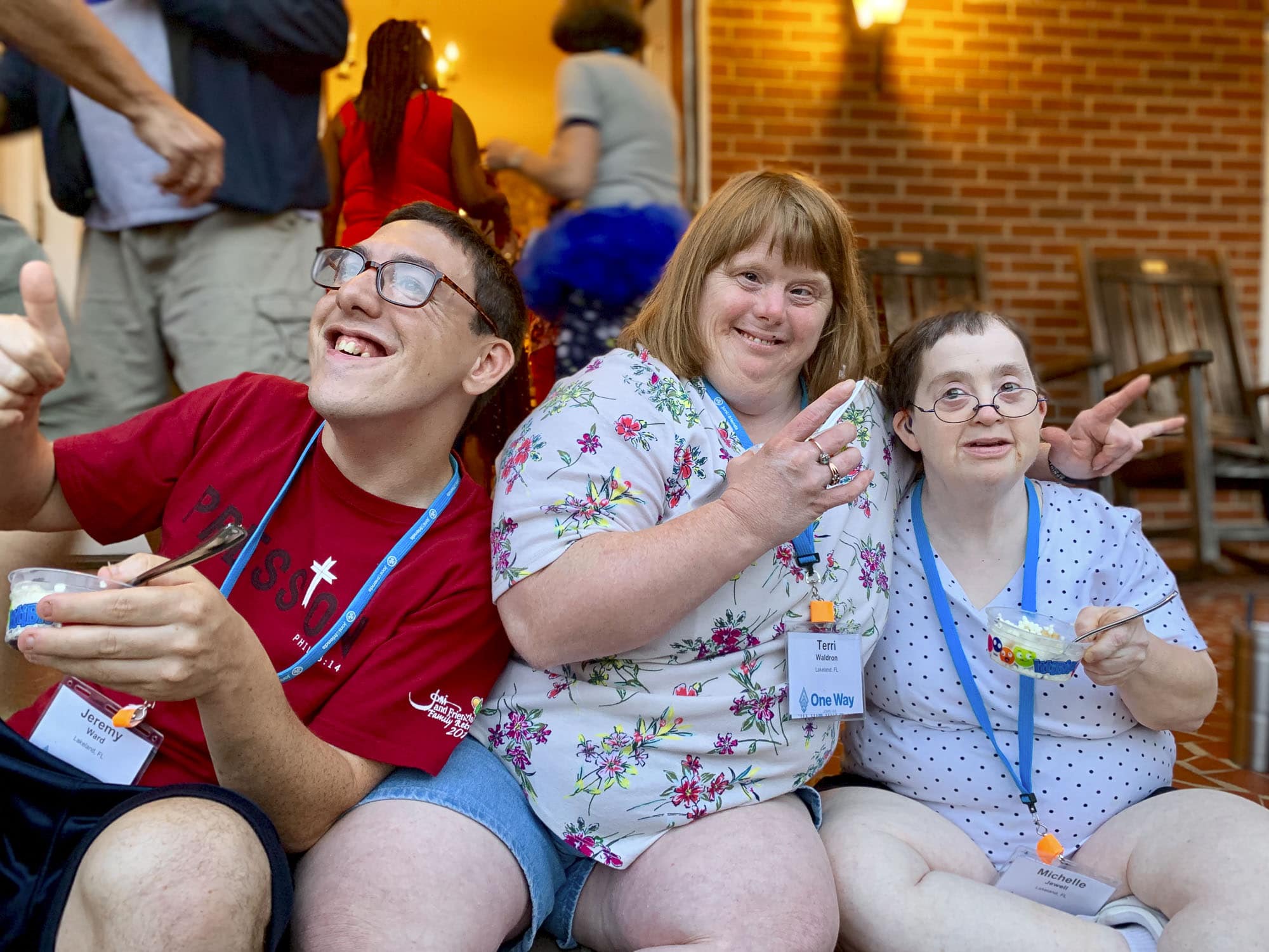Three adult-aged people who have developmental disabilities, one man on the right and two women on the left who are hugging each other and smiling at the camera.