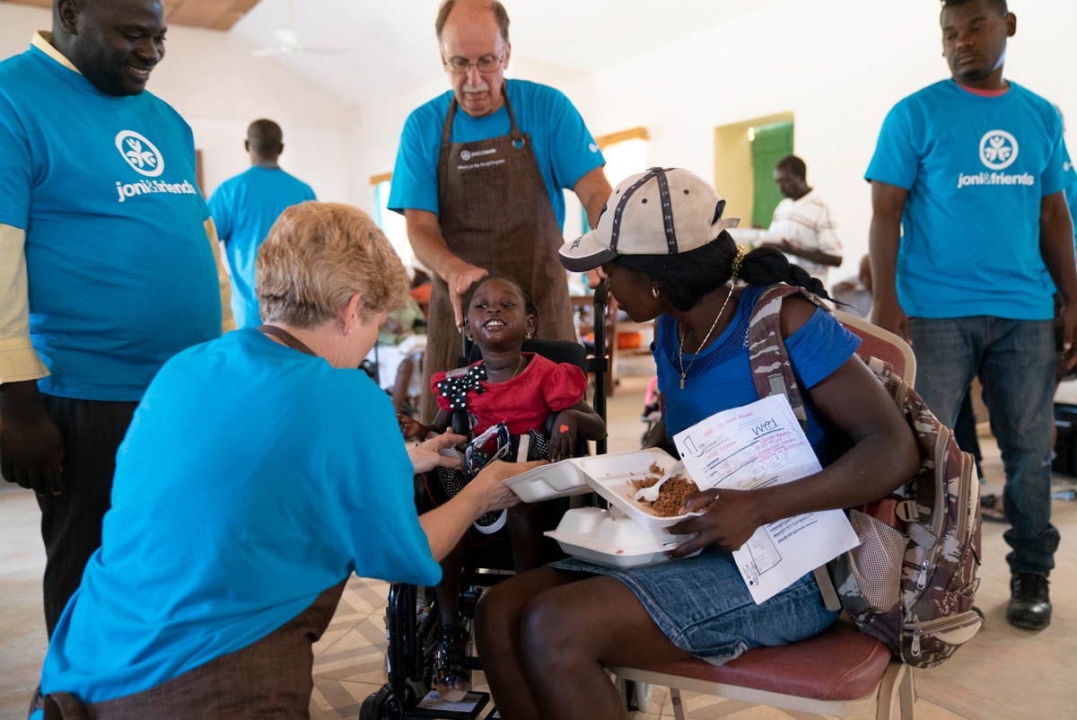Woodlove in her new wheelchair smiling with four Joni and Friends volunteers around her helping to adjust her wheelchair. Her mom is sitting beside her and smiling with two containers of food in her lap.