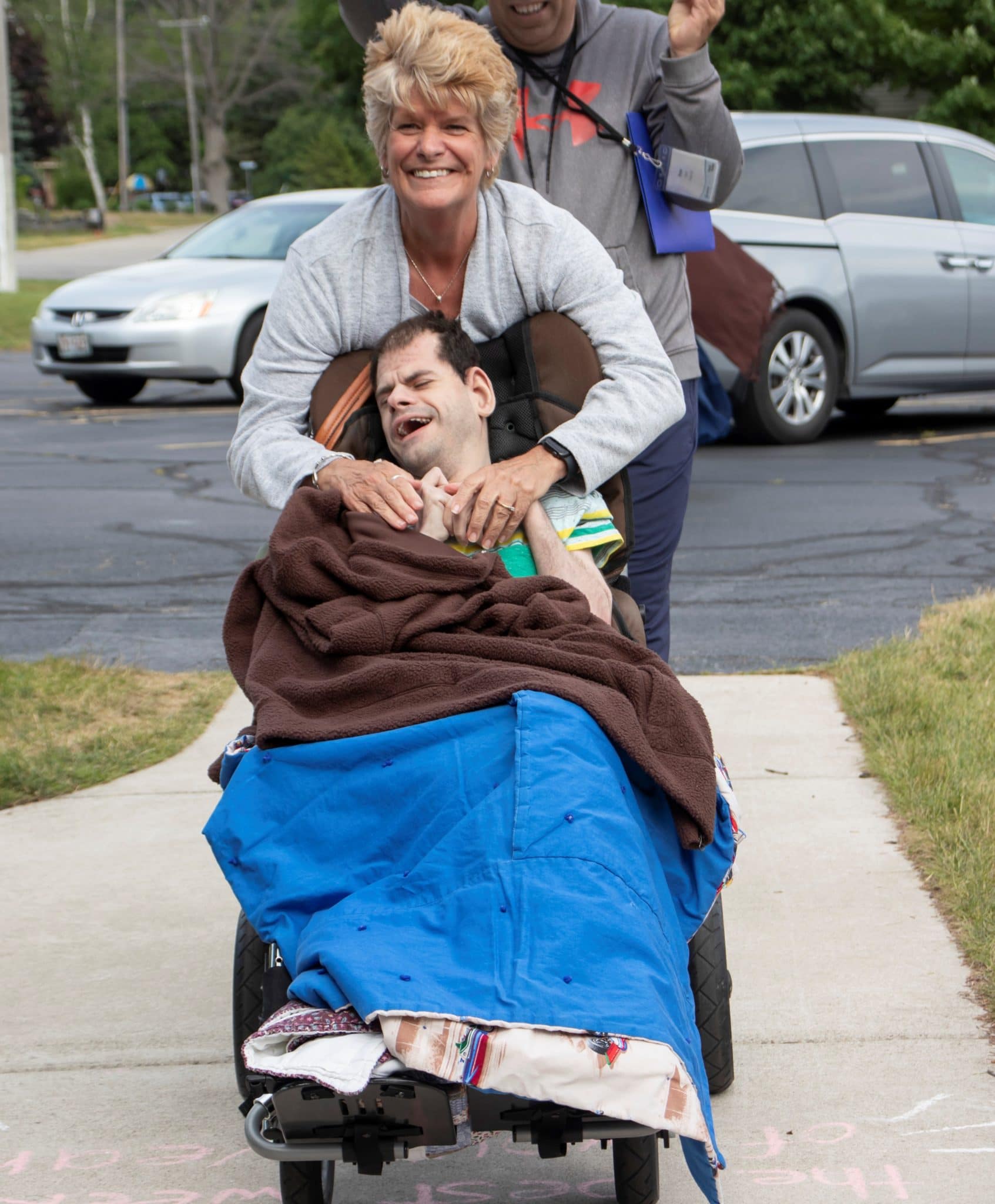 Buddy's mom, Sheila, smiling wide as she pushes Buddy's wheelchair towards the retreat center. Buddy is also smiling and holding his mom's hands, he has a couple blankets over his lap.