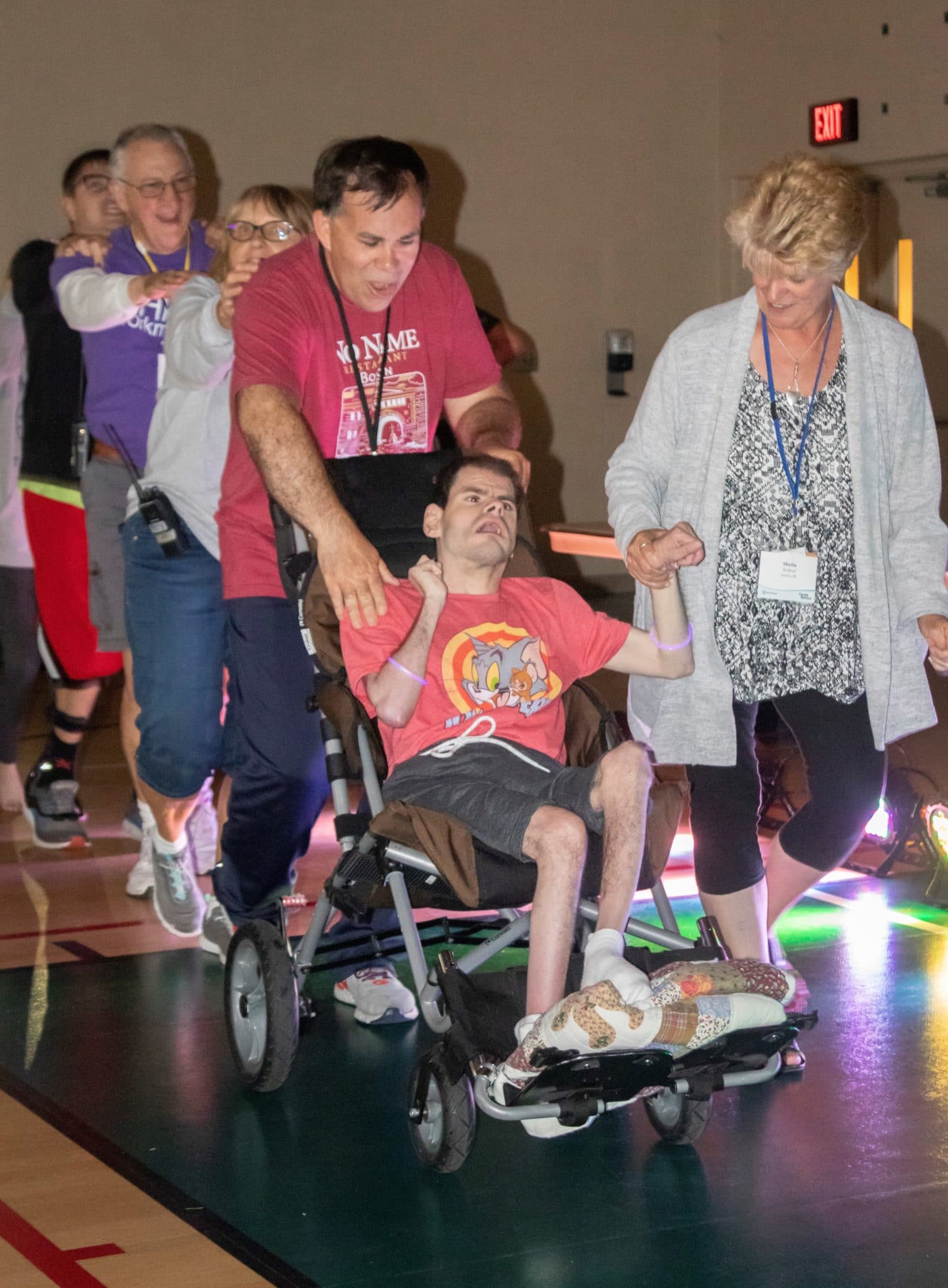Buddy being pushed in his wheelchair at the front of a conga line, his mom, Sheila, looking down at him, smiling and holding his hand.