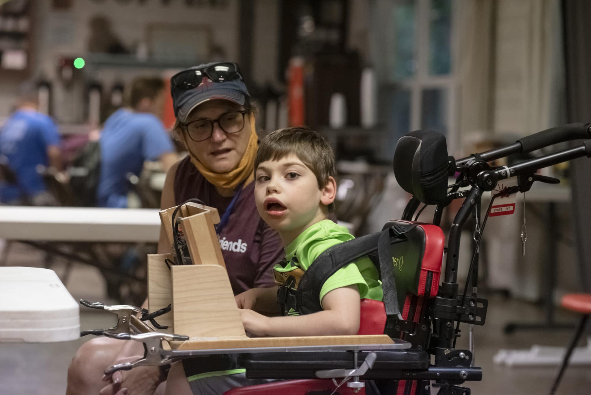 Qorban sits in his wheelchair using his speech device to communicate with his Family Retreat volunteer sitting next to him on his right side and spending the day with him. Qorban is looking at the camera while his volunteer looks at him.