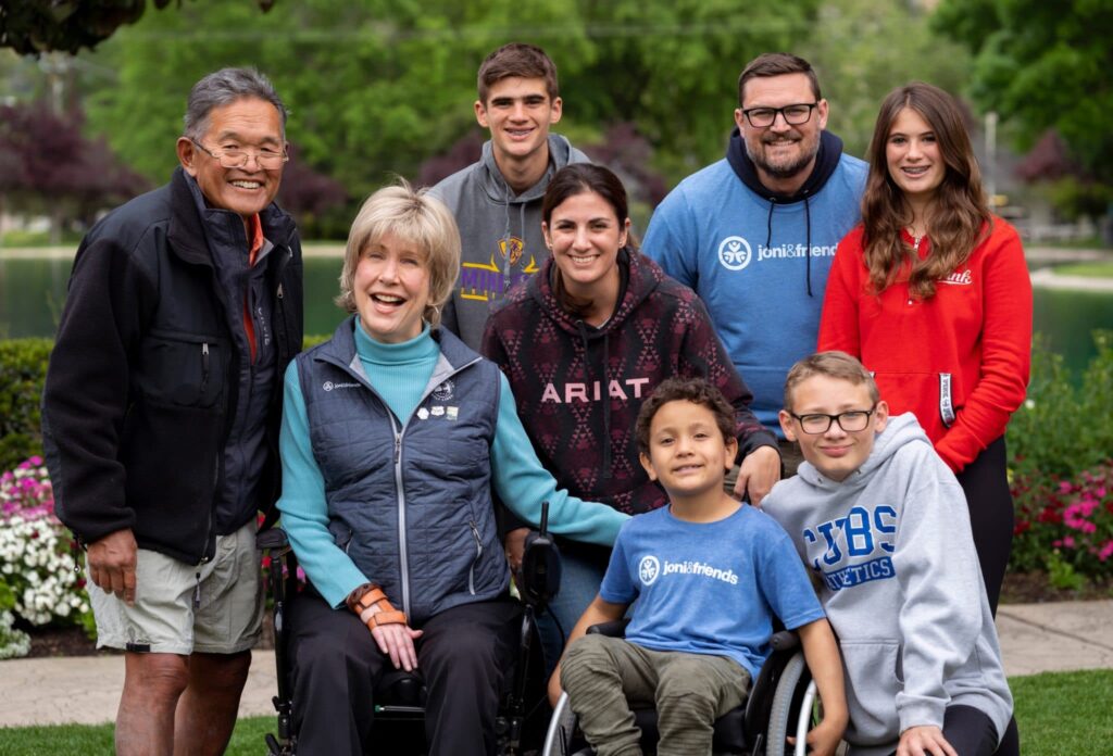 Ken Tada, Joni Eareckson Tada, Logan, Luke and Sophie, Ryan, Christen and Leo in his wheelchair posing together in front of a pond and smiling at the camera.