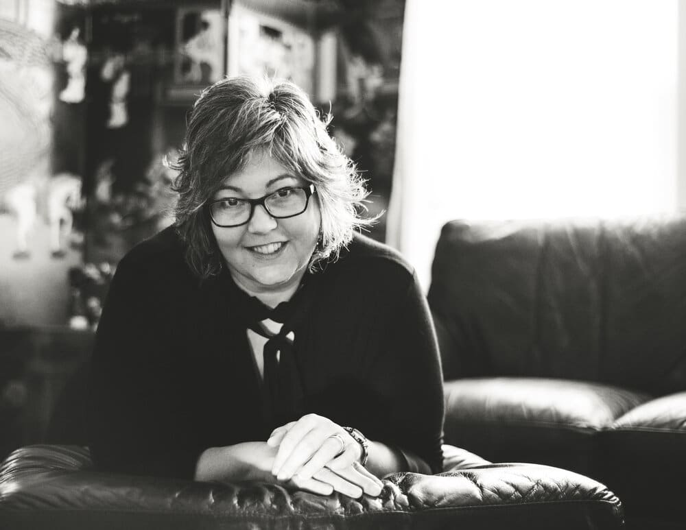 A black and white photo of Danika Deva leaning over a leather ottoman. She has glasses and short hair, with her right hand over her left as she smiles at the camera.