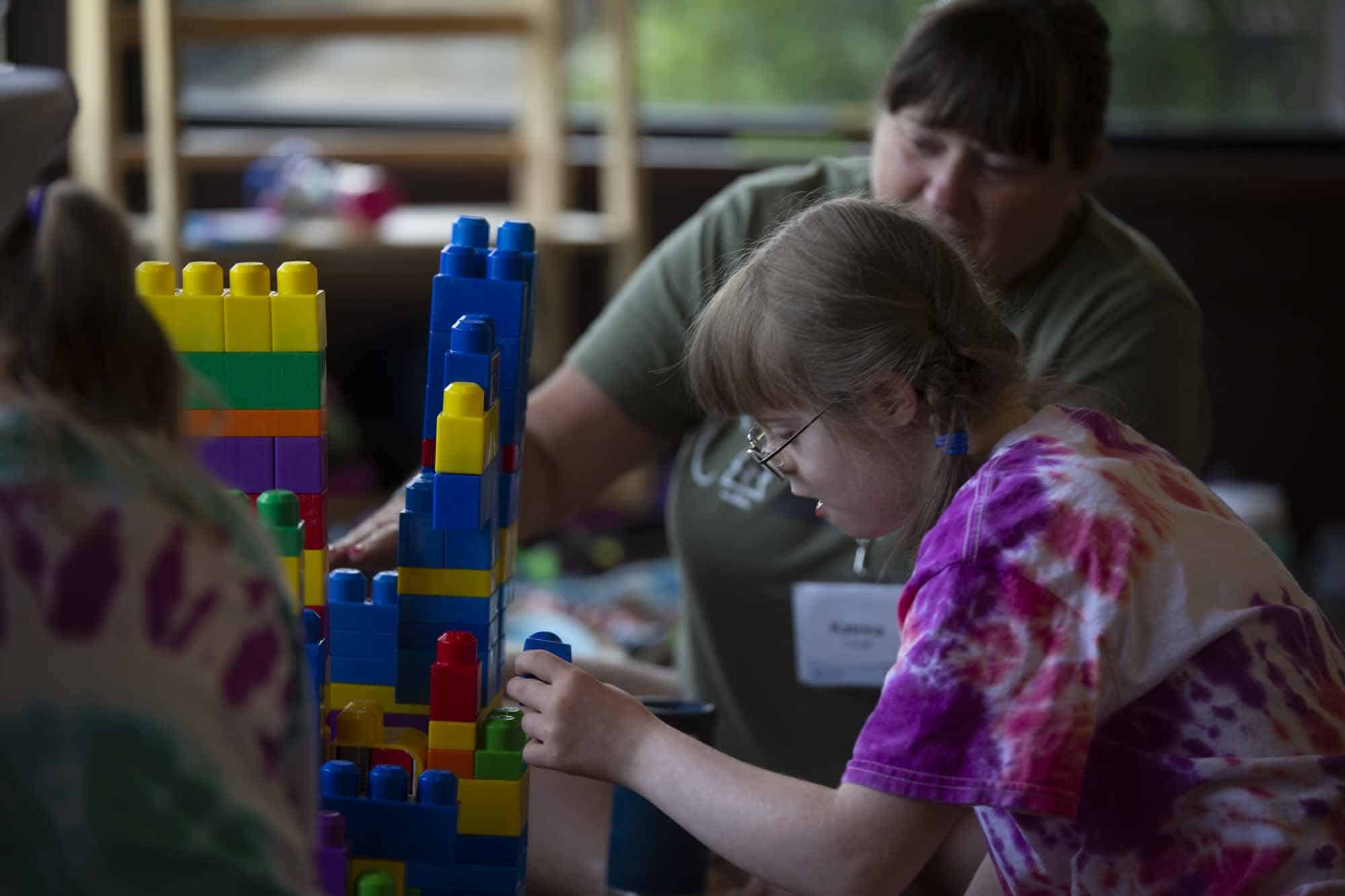 A young girl with down syndrome playing Legos with gold reading glasses on and a tie-dye shirt, an older woman sitting next to her playing Legos along side her.