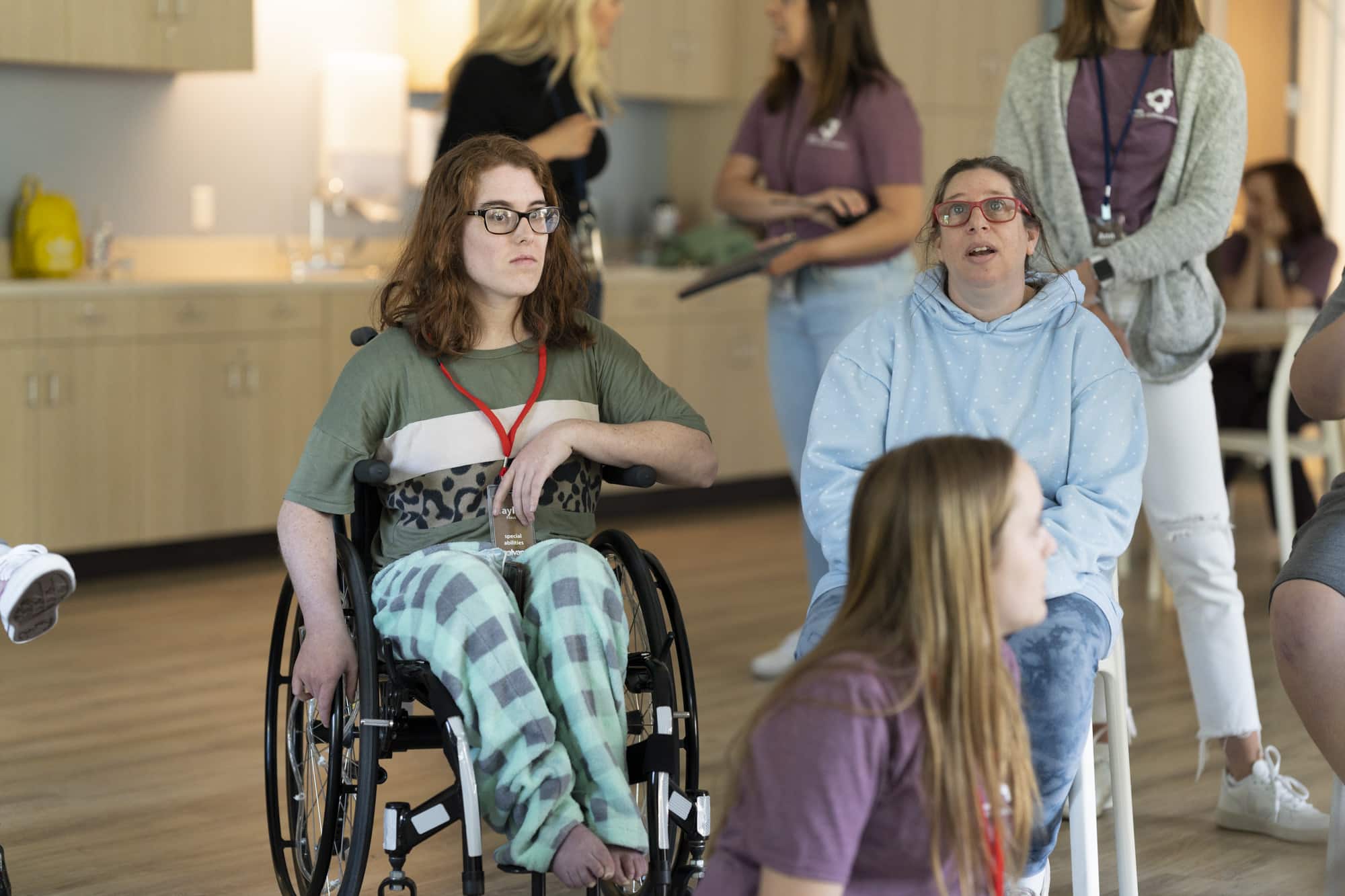 A young woman seated in her wheelchair looking off into the distance and another young woman seated next to her in a chair who appears to have a cognitive disability.