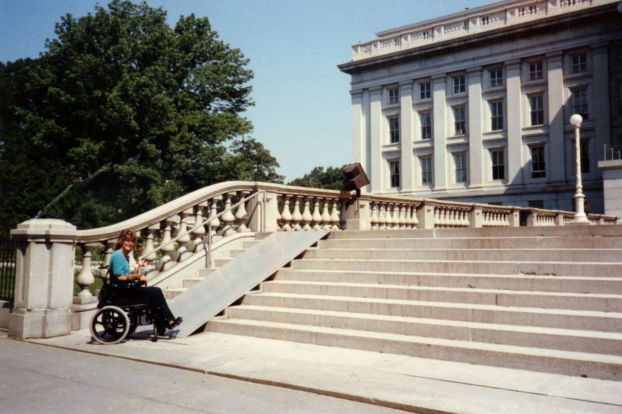 Joni seated in her wheelchair at the bottom of a set of stairs in front of a large historical building, a recently installed ramp in front of her.