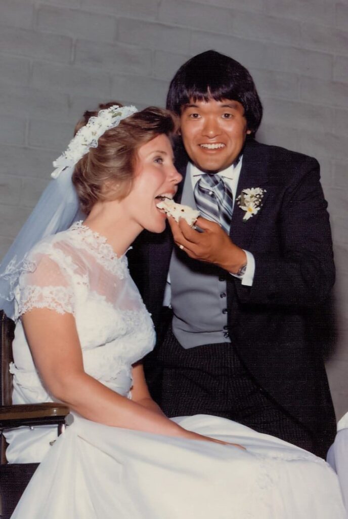 Joni and Ken on their wedding day. Ken is feeding Joni a slice of wedding cake while she sits in her wheelchair and wedding dress.